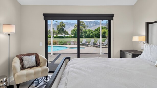 Quarto de hotel com cama, cadeira, mesa de cabeceira, luminária e vista para uma piscina e montanhas.