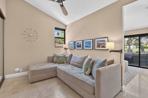 A modern living room with a sectional sofa, a ceiling fan, a wall clock, framed pictures, and a view through a glass door. - Elegant South-end (Less Wind) 3BD/3BA Right On The Fairway! (Palm Springs)