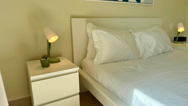 Una cama bien hecha con sábanas blancas, una mesita de noche con lámpara y una planta en maceta.