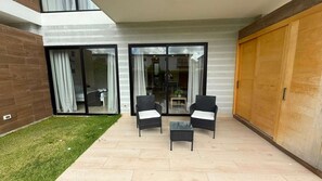 A modern outdoor seating area with two chairs and a small table, a sliding glass door leading to a room, and a wooden door on the right. - NOK Stylish 1BR in Ventura By Vistacana Rentals (Punta Cana)
