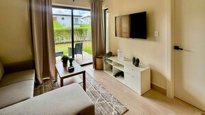 Una sala de estar moderna con un sofá, una mesa de centro, un televisor montado en la pared y un ventanal corredizo que da a un espacio exterior.