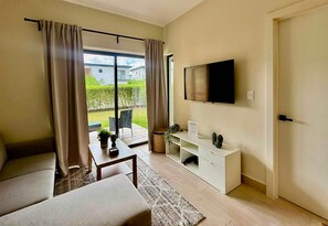 A modern living room with a sofa, a coffee table, a TV mounted on the wall, and a sliding glass door leading to an outdoor area. - NOK Stylish 1BR in Ventura By Vistacana Rentals (Punta Cana)