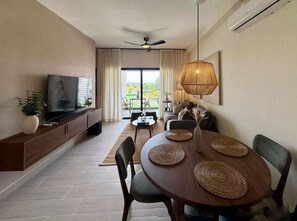 Ein modernes Wohnzimmer mit Flachbildfernseher, einem Esstisch mit gewebten Platzsets, einem Deckenventilator und einem großen Fenster mit Vorhängen.
