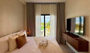 Ein Hotelzimmer mit Bett, Fernseher, einem Balkon mit Vorhängen und Blick auf Grünflächen.