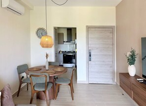 Et spiseområde med et rundt bord, fire stole, en hængende lampe og et køkken i baggrunden.