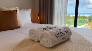 Et hotelværelse med en seng, to rullede håndklæder med teksten 'VISTACANA', et vindue med udsigt over en golfbane og et natlys på væggen.