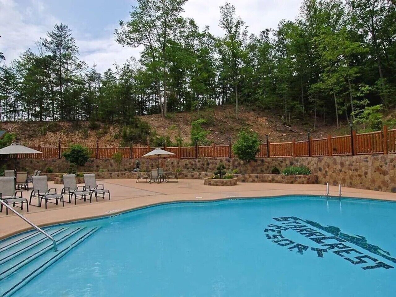 A swimming pool with lounge chairs and umbrellas, surrounded by a wooden fence and trees.