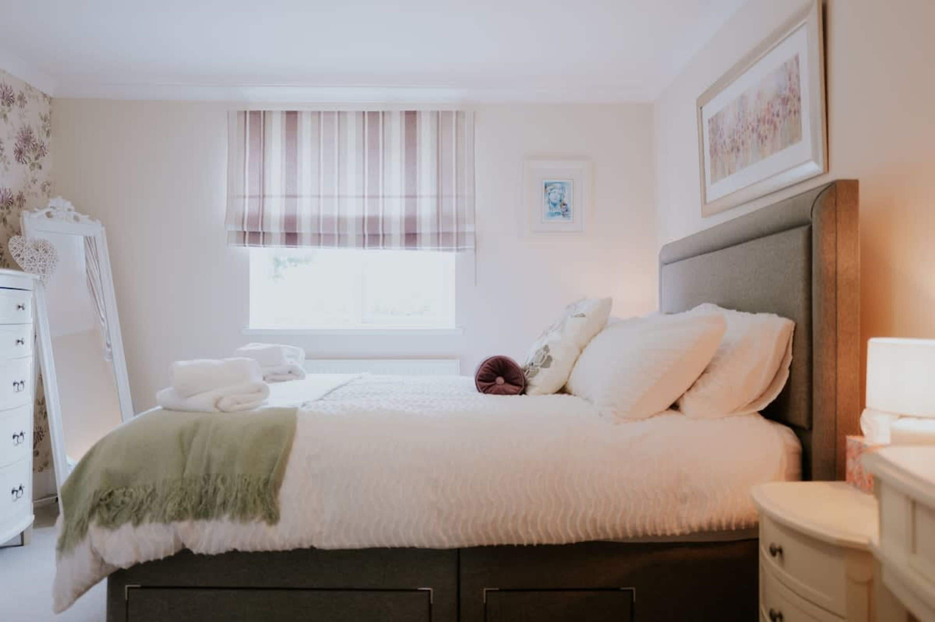 Un lit bien fait, avec des oreillers et une couverture, une table de chevet, une commode, une fenêtre avec des rideaux et un tableau encadré au mur.
