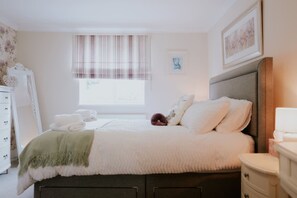 A neatly made bed with pillows and a blanket, a bedside table, a dresser, a window with curtains, and a framed picture on the wall. - Perfect Booking 3 Bed Home With Beautiful Garden (Ashford)