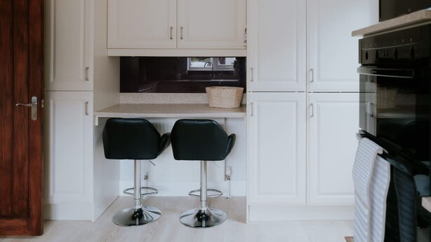 A compact kitchen with white cabinets, a black countertop, and two black bar stools. - Perfect Booking 3 Bed Home With Beautiful Garden (Ashford)