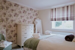 Une chambre avec un papier peint à motifs floraux, une commode blanche, un lit recouvert d’un plaid vert et une fenêtre agrémentée d’une lambrequin à rayures.