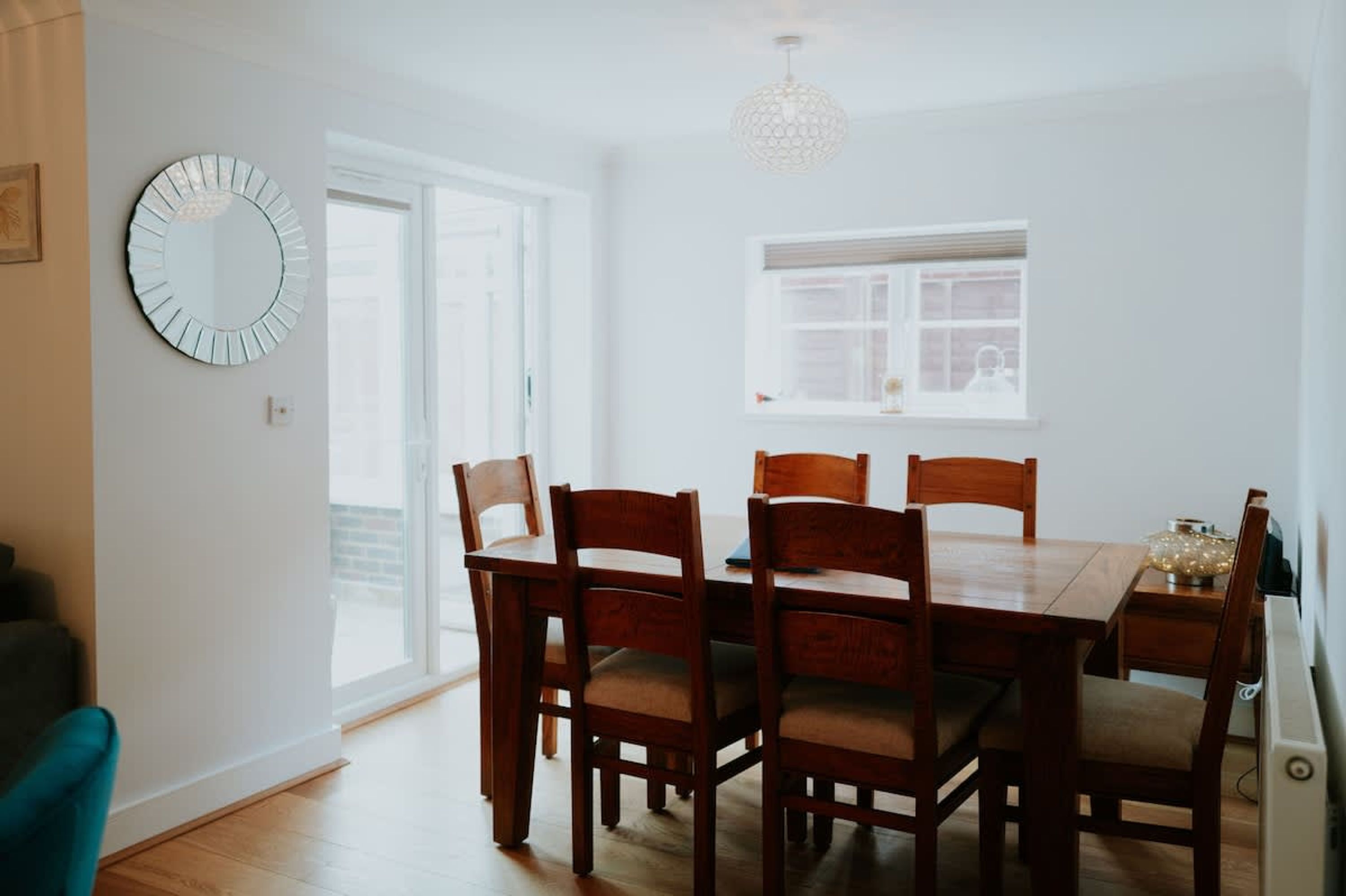 A dining area with a wooden table and chairs, a round mirror on the wall, and a window with blinds.