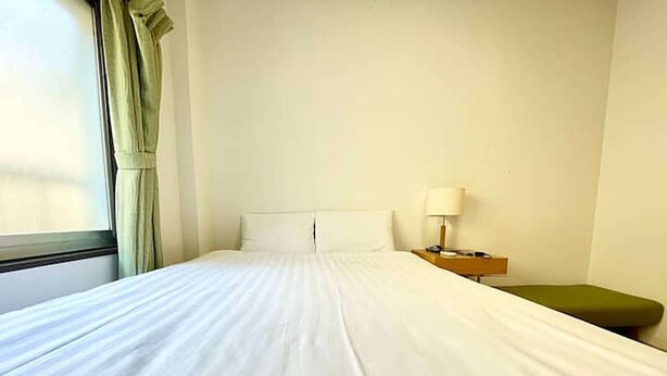 Quarto de hotel com uma cama grande, uma mesa de cabeceira com abajur e uma janela com cortinas.