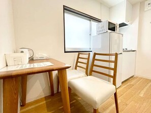 A small kitchenette with a microwave, a refrigerator, and a window with blinds. - LunaStay Apartments - 103 (東京)