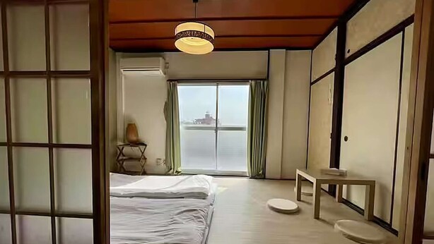 A traditional Japanese-style room with tatami flooring, a bed, a small table, and a view of the sea through a sliding door. - LunaStay Apartments - 301 (東京)