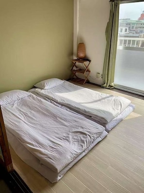 Ein Zimmer mit zwei Betten, einem kleinen Tisch mit einer Lampe und einem Fenster mit Blick auf Gebäude.