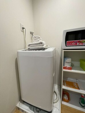 Une machine à laver blanche, sur laquelle est empilée une pile de serviettes blanches, une prise de courant et une étagère contenant divers objets, notamment une boîte étiquetée « Penguin » et une bouteille de produit de nettoyage.