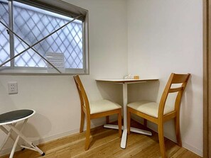 Ein Eckzimmer mit einem kleinen Tisch und zwei Holzsesseln, einem Fenster mit einem quadratischen Rollo sowie einer Steckdose an der Wand.
