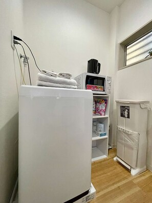 Eine kompakte Küche mit einem weißen Kühlschrank, einer Mikrowelle und einem kleinen Regal mit verpackten Lebensmitteln.