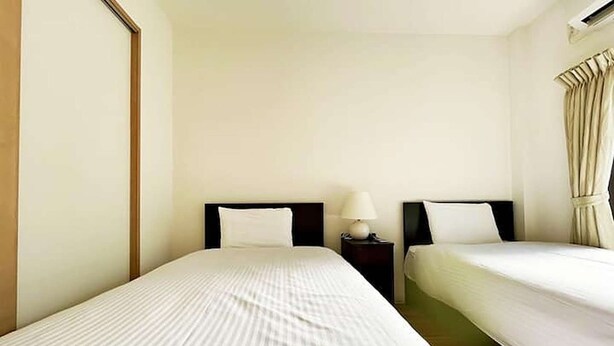 Quarto de hotel com duas camas de solteiro, uma mesinha de cabeceira com abajur e uma janela com cortinas.