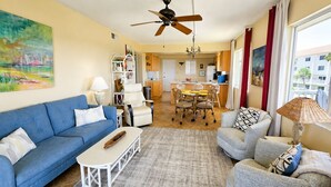 A living room with a blue sofa, a grey armchair, a coffee table, and a dining area with a table and chairs. - Beachfront Ocean-Views Condo, Balcony! Pool, BBQ (St. Augustine Beach)