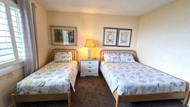 A bedroom with two single beds, a nightstand with a lamp, and framed pictures on the wall. - Beachfront Ocean-Views Condo, Balcony! Pool, BBQ (St. Augustine Beach)