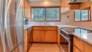 A modern kitchen with wooden cabinets, a stainless steel refrigerator, and a large window providing a view of trees. - Cozy Sunriver Retreat, 6 SHARC, AC, Loft & Wood Stove (Sunriver)