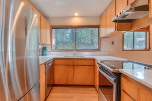 A modern kitchen with wooden cabinets, a stainless steel refrigerator, and a large window providing a view of trees. - Cozy Sunriver Retreat, 6 SHARC, AC, Loft & Wood Stove (Sunriver)
