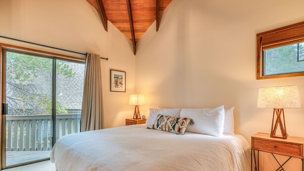 A bedroom with a large bed, two bedside lamps, a window with a view of a wooden deck, and a ceiling with exposed beams. - Cozy Sunriver Retreat, 6 SHARC, AC, Loft & Wood Stove (Sunriver)