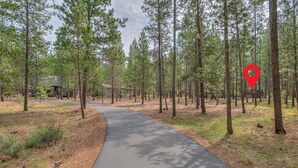 A forested area with a paved path, tall pine trees, and a building in the background. - Cozy Sunriver Retreat, 6 SHARC, AC, Loft & Wood Stove (Sunriver)
