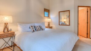 A bedroom with a bed, two bedside tables with lamps, a bedside clock, a window with blinds, a mirror on the wall, and a wooden door. - Cozy Sunriver Retreat, 6 SHARC, AC, Loft & Wood Stove (Sunriver)