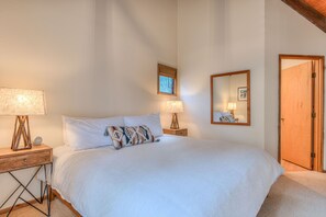 A bedroom with a bed, two bedside tables with lamps, a bedside clock, a window with blinds, a mirror on the wall, and a wooden door. - Cozy Sunriver Retreat, 6 SHARC, AC, Loft & Wood Stove (Sunriver)