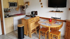 Eine kompakte Küche mit Mikrowelle, Spüle und Holzausbau. Ein Esstisch mit Stühlen und ein schwarzer Mülleimer sind vorhanden. Ein Flachbildfernseher ist an der Wand montiert.
