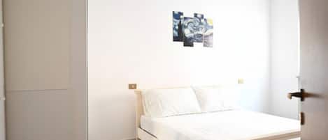 A bedroom with a bed, white walls, and a small painting on the wall.