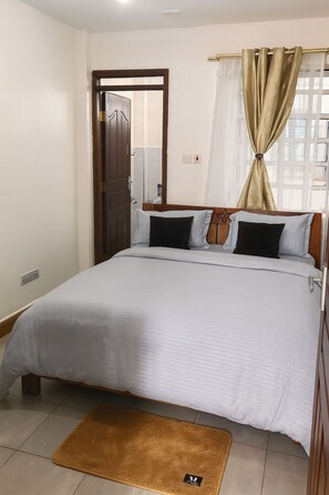 A bedroom with a bed, a wooden door, a window with curtains, and a small rug. - 2-bedroom in secure leafy area Conveniently close to a coffee shop & restaurants (Nairobi)