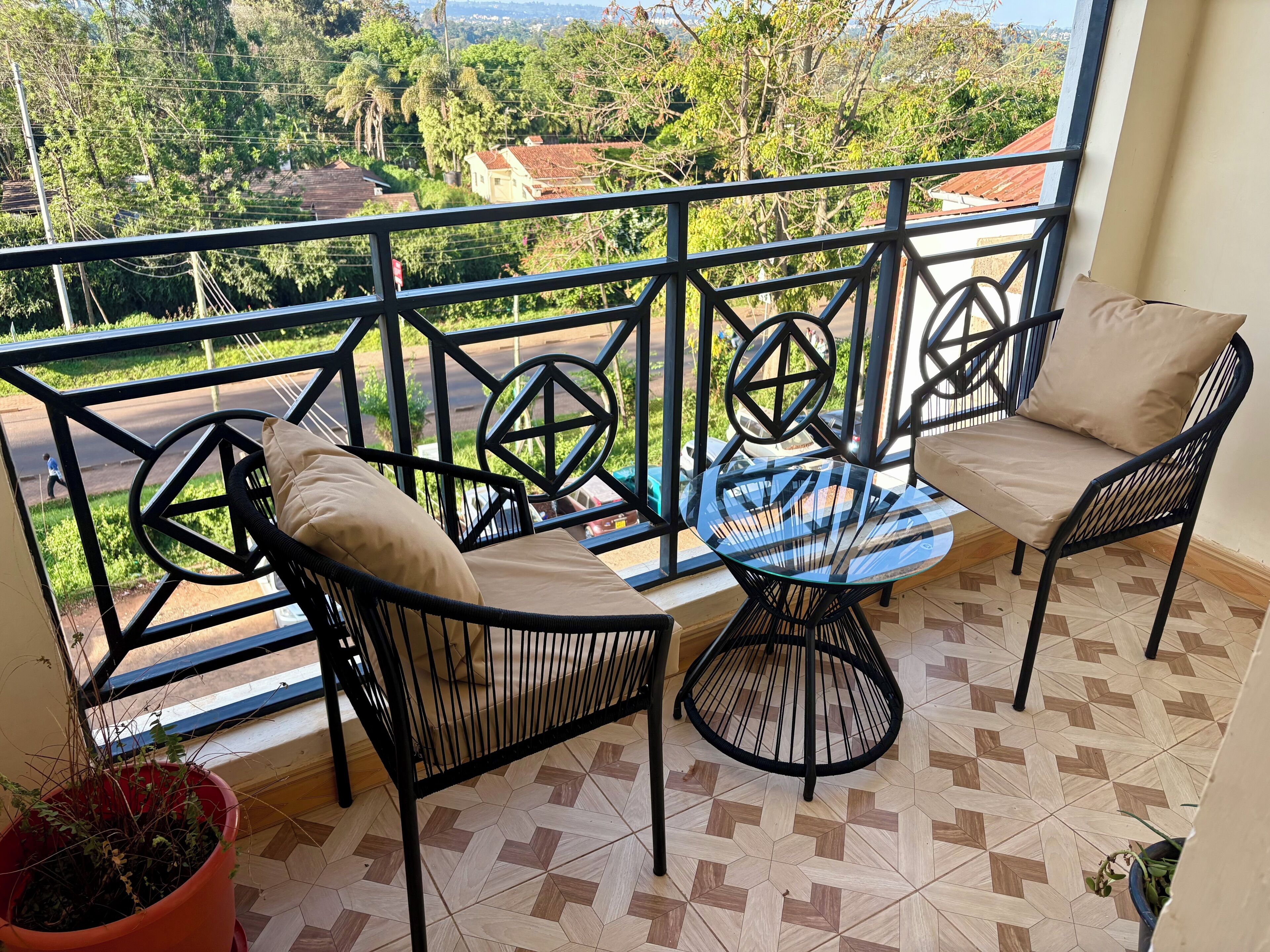 A balcony with a glass table and two chairs, a potted plant, and a view of a road and greenery.