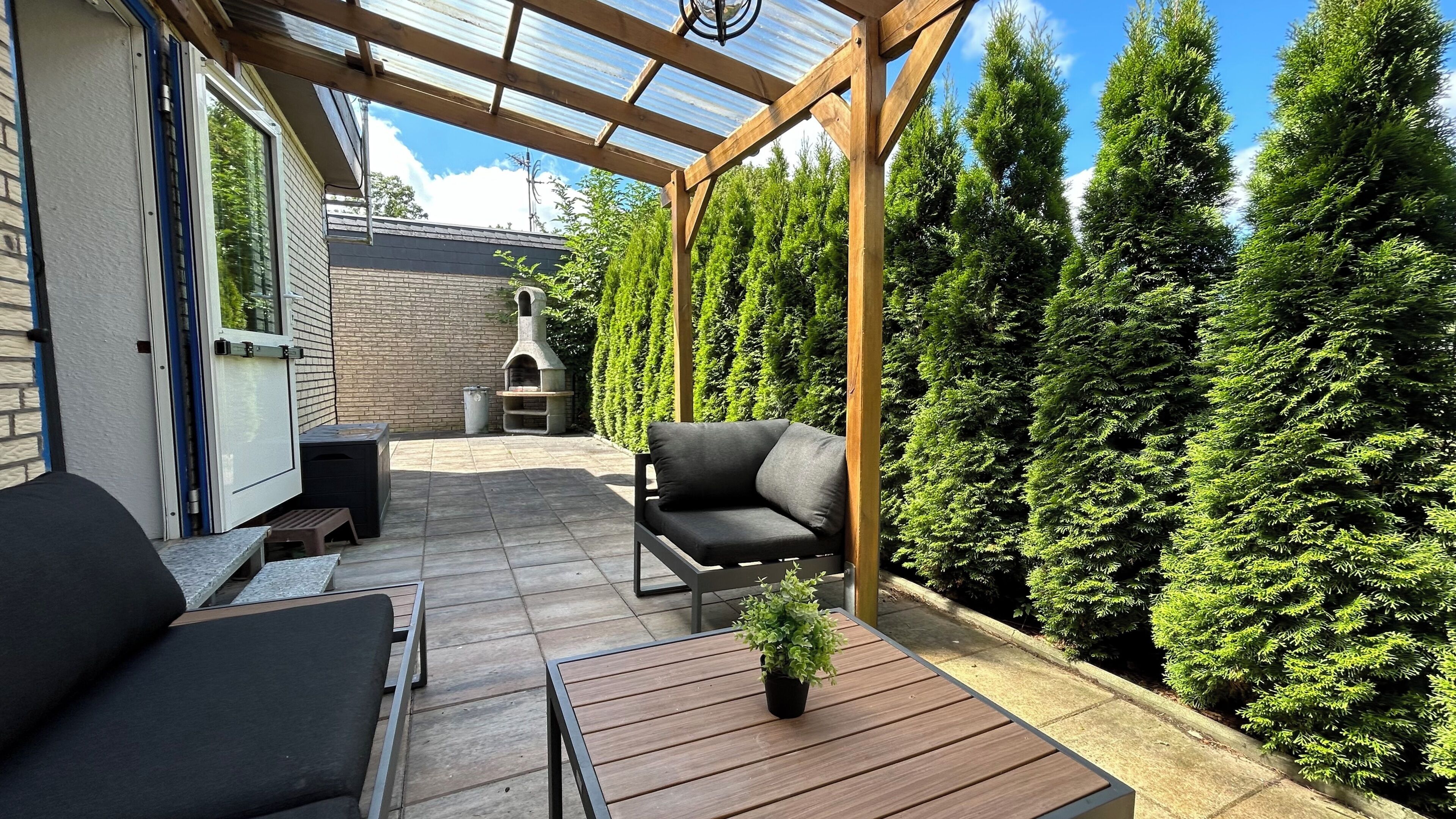 Een overdekt terras met een houten pergola, een stenen openhaard, zitplaatsen buiten en uitzicht op hoge altijdgroene bomen.