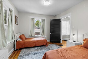 A bedroom with two beds, a window with curtains, a door, a bedside table, and a rug.