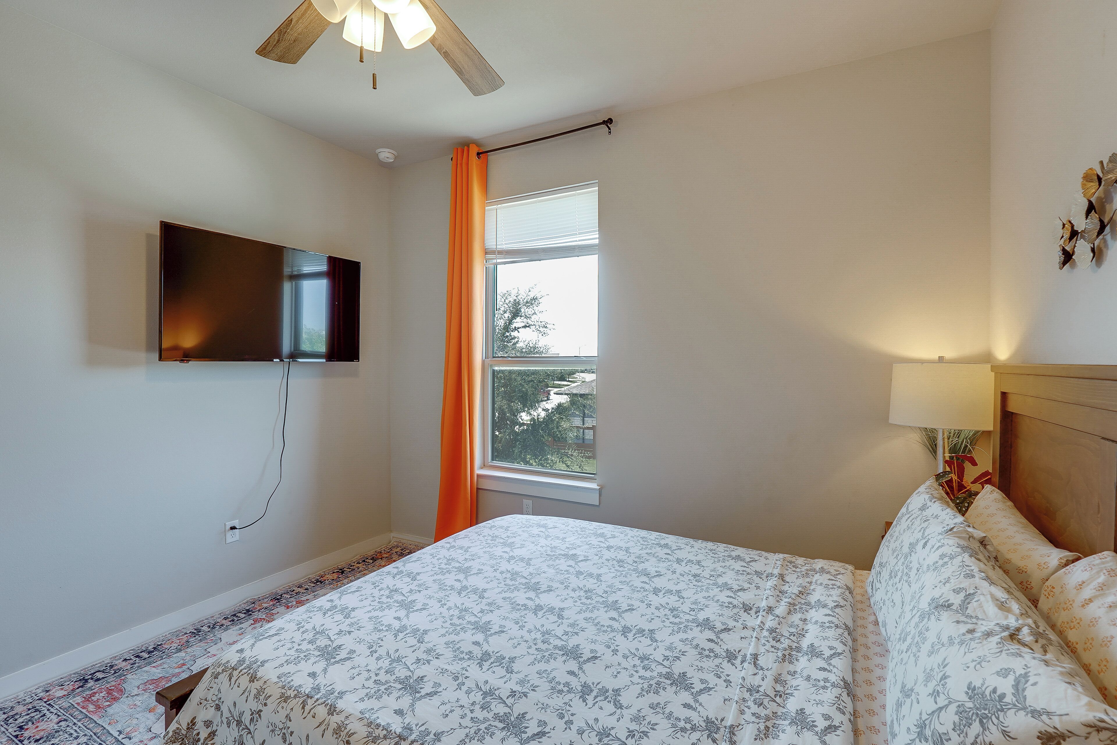Une chambre à coucher avec un lit, un téléviseur fixé au mur, une fenêtre avec des rideaux et un ventilateur de plafond.