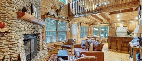 A cabin interior with a stone fireplace, wooden walls, a loft with a railing, and a kitchen area.