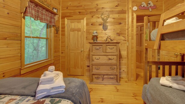 A wooden cabin interior with two beds, a dresser with drawers, a window with a view of trees, and a wall-mounted lamp. - Hot Tub & Game Room! Wooded Retreat in Blue Ridge (Blue Ridge)