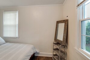 A bedroom with a bed, a window with blinds, a wall-mounted mirror, and a metal shelf.
