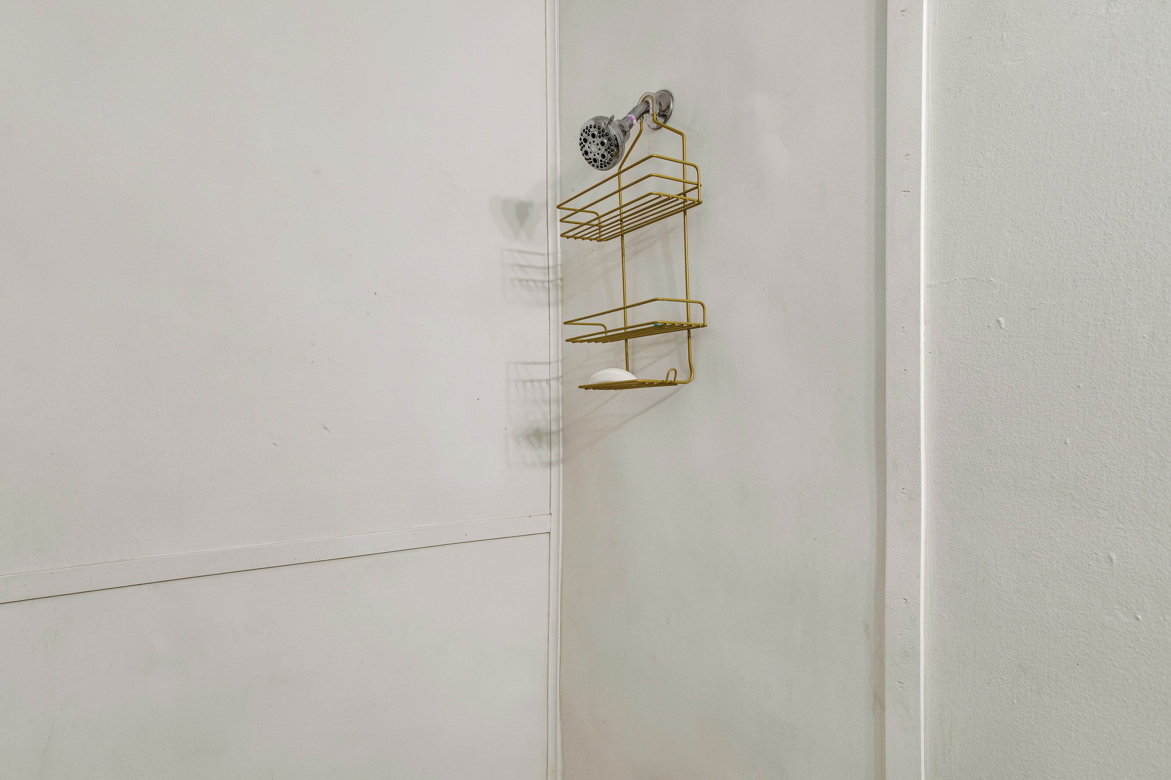 A bathroom with a white wall, a shower, and a gold wire shelf unit.