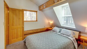 A bedroom with a sloped ceiling, a wooden door, a window, a bedside table with a lamp, and a bed with patterned bedding. - Real Log Cabin in Flagstaff With Forest Views (Flagstaff)