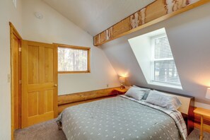 Ein Schlafzimmer mit schrägverdeckter Decke, einer Holztür, einem Fenster, einem Nachttisch mit Lampe und einem Bett mit gemusterten Betttextilien.