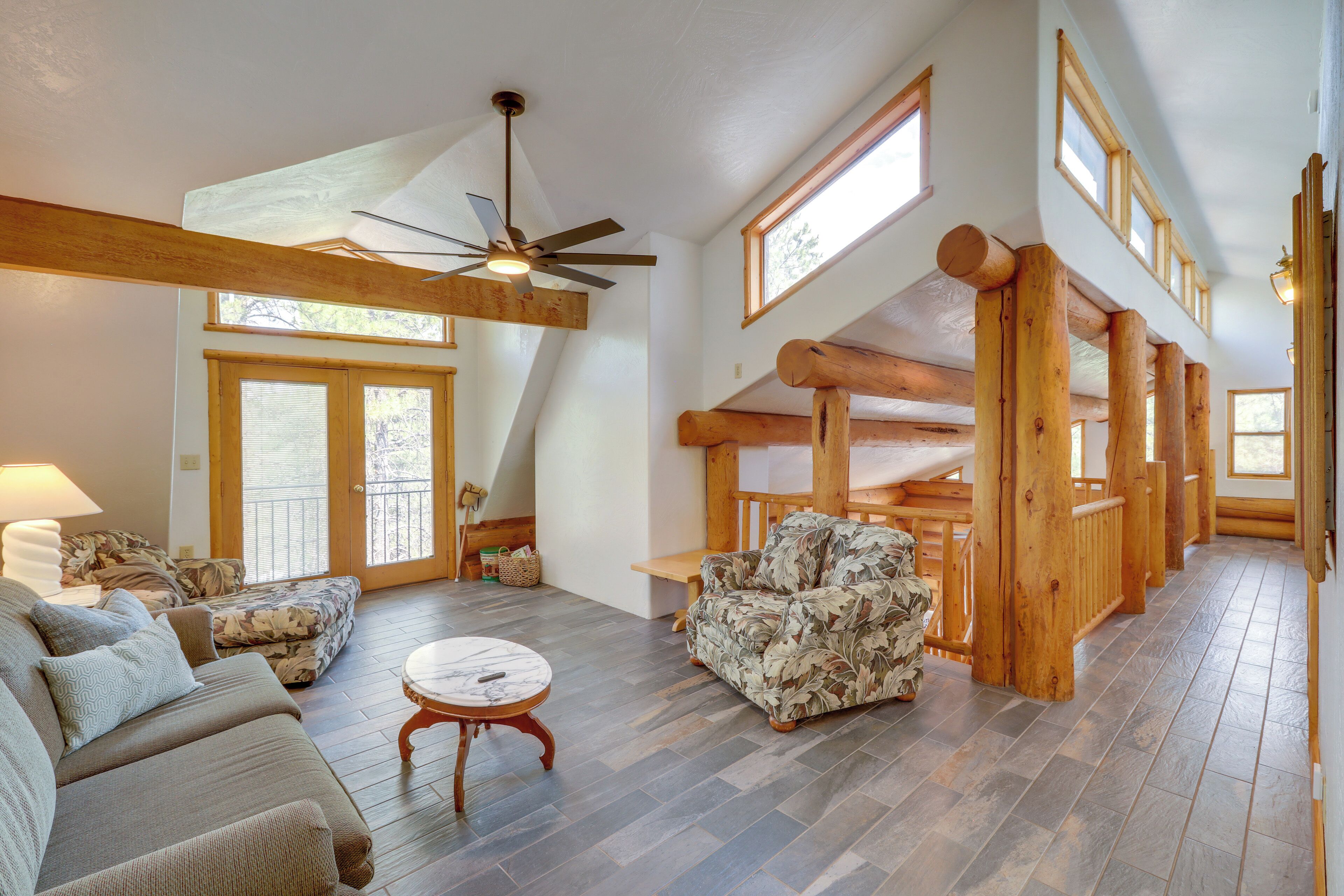 A spacious living area with a high ceiling, wooden beams, a ceiling fan, and a large window.