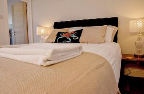 A neatly made bed with a folded white towel, a black pillow with a colorful design, and two bedside lamps on a wooden nightstand. - Relax at Pebble House for a seaside break. (Whitstable)