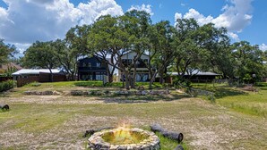 A stone fire pit with logs, surrounded by a grassy area with trees and a two-story house in the background. - Walk to Shore! Family-friendly Canyon Lake Retreat (Canyon Lake)