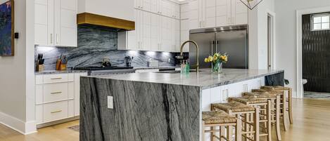 A modern kitchen with white cabinets, a marble island, a stainless steel refrigerator, and a unique pendant light.