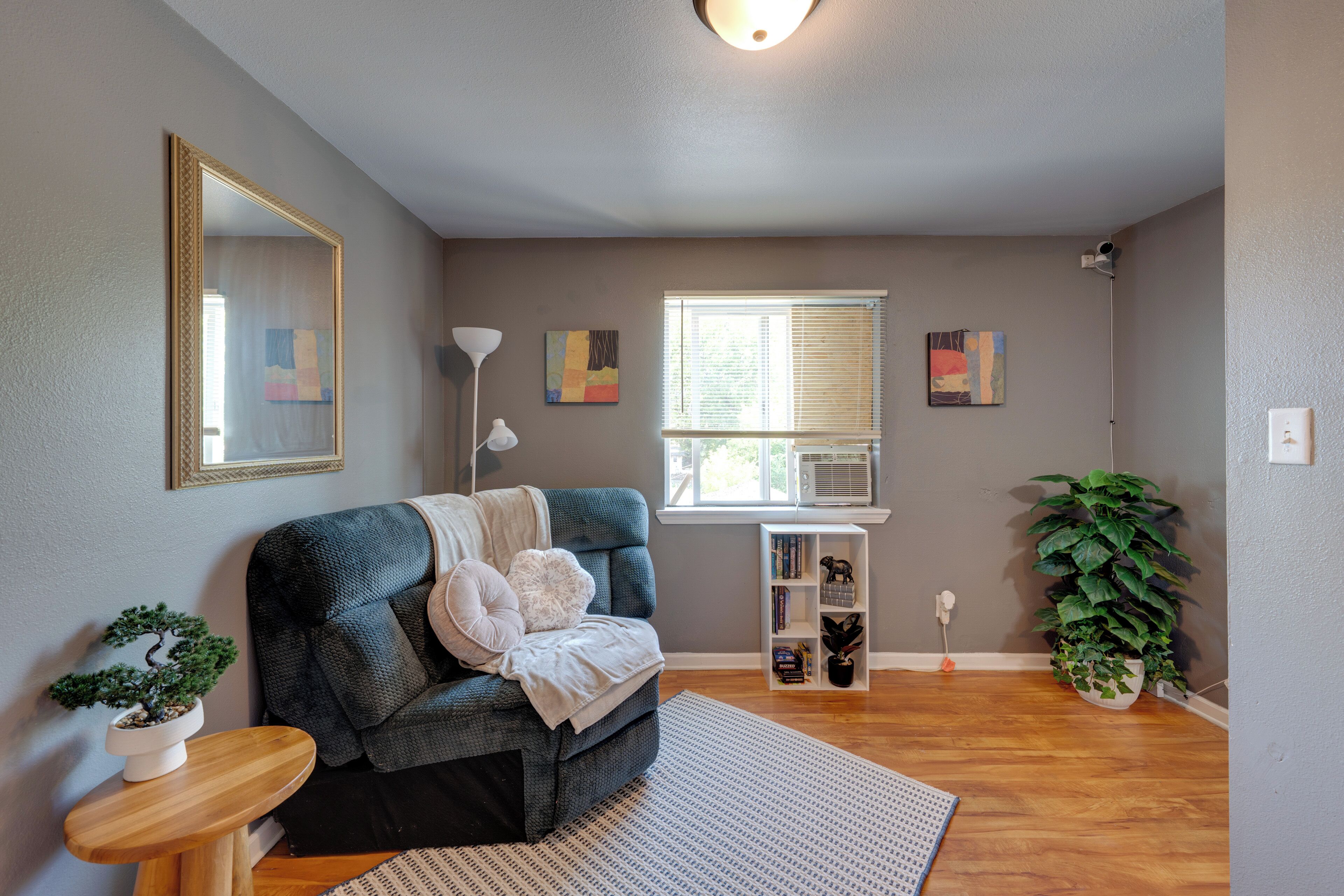 A living room with a sofa, a side table with a plant, a standing lamp, and a window with blinds.